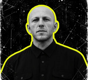 DVS1 wearing a dark button-up shirt, in a black-and-white portrait with a bright yellow outline.