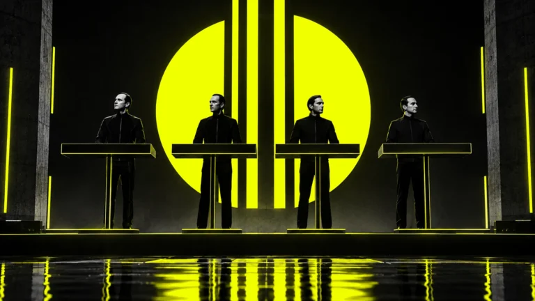 Four anonymous electronic musicians behind synthesizer consoles on a dark industrial stage with acid-yellow geometric lighting.