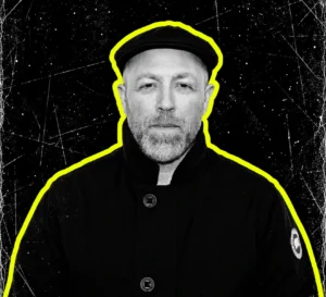 Ben Sims wearing a flat cap and dark coat, in a black-and-white portrait with a yellow outline.